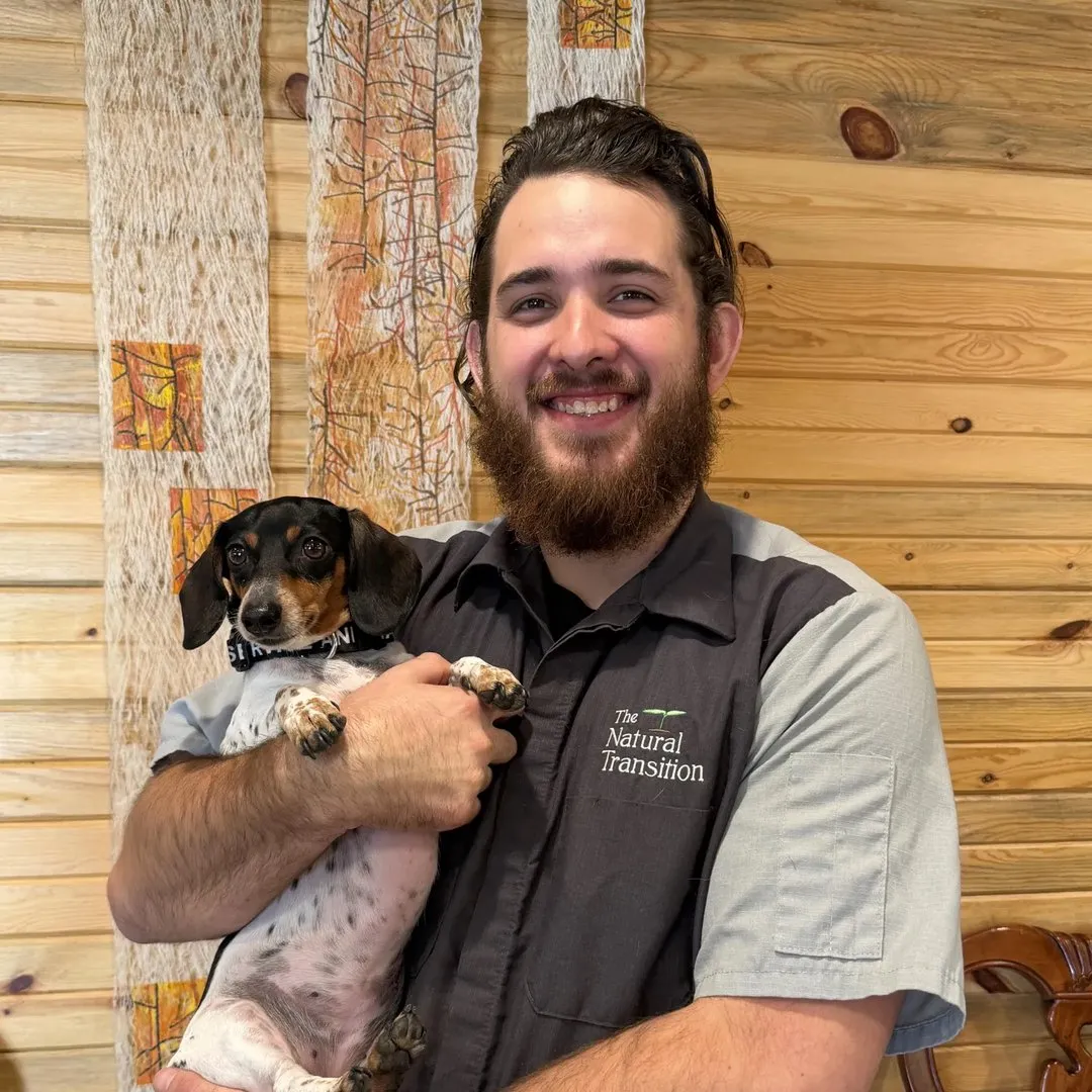 Ragen Hale smiling in a Natural Transition work shirt, holding a small black-and-white dachshund, against a warm wood-slat wall.