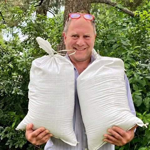 Dan Ziskin, co-owner of The Natural Funeral, smiling under a tree and holding two full 25-pound bags of soil